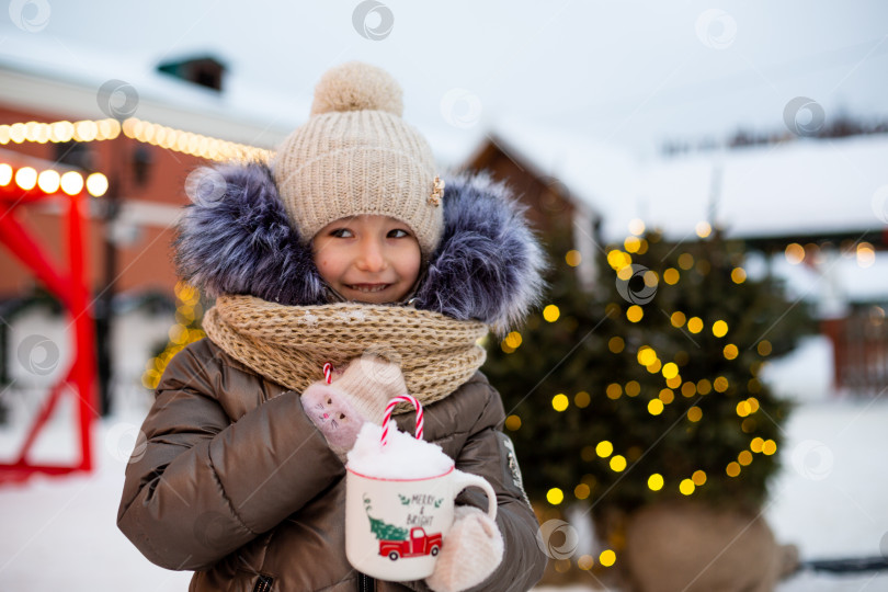 Скачать Девушка с кружкой со снегом, леденцовой палочкой и надписью Merry and Bright в руках на улице в теплой одежде зимой на праздничном базаре. Гирлянды сказочных огней украсили снежный городок к новому году. Рождество фотосток Ozero