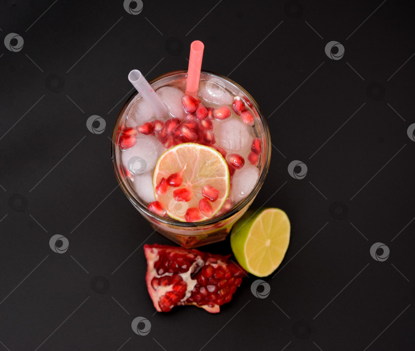 Скачать Стакан фруктового сока со льдом и фруктовыми косточками на черном фоне, рядом со спелым гранатом и лаймом. фотосток Ozero