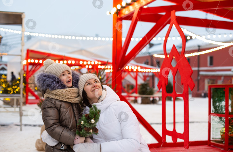 Скачать Мама и дочь на Рождество на открытом воздухе в теплой одежде зимой на праздничном рынке. Сказочные огни и гирлянды украсили снежный городок к новому году фотосток Ozero