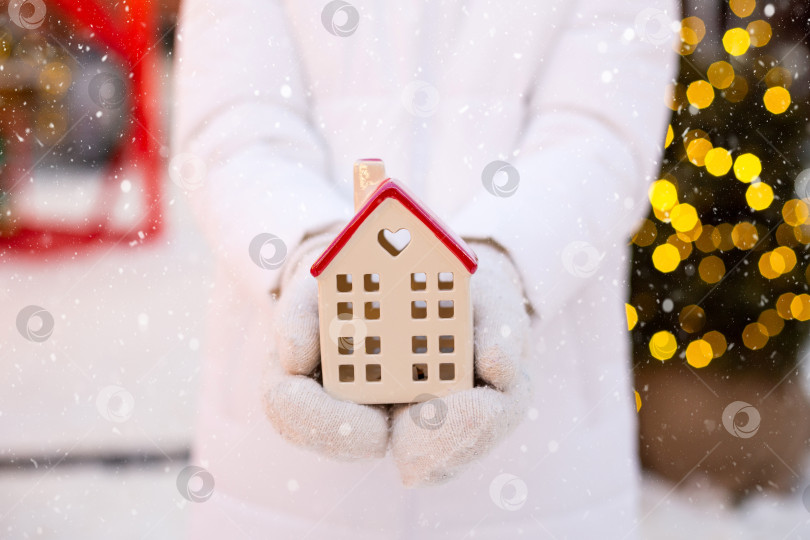 Скачать Дом, миниатюрный коттедж в руках женщины, одетой в варежки и теплую одежду на улице в снегу. Сделки с недвижимостью, покупка, строительство, переезд, ипотека. Бронирование уютного дома на Рождество, новый год фотосток Ozero