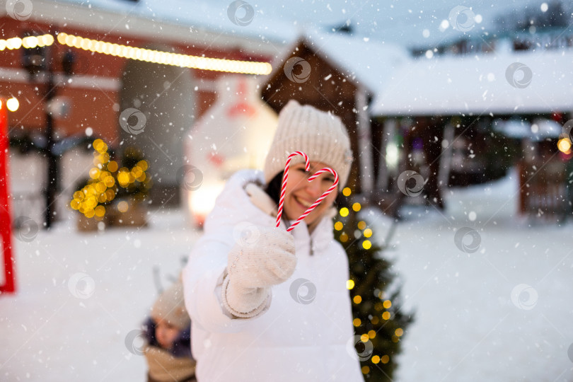Скачать Женщина держит в руках леденцовый тростник в форме сердца на открытом воздухе в белой теплой одежде на зимнем праздничном рынке. Гирлянды сказочных огней украсили снежный городок к новому году. Рождественское настроение фотосток Ozero