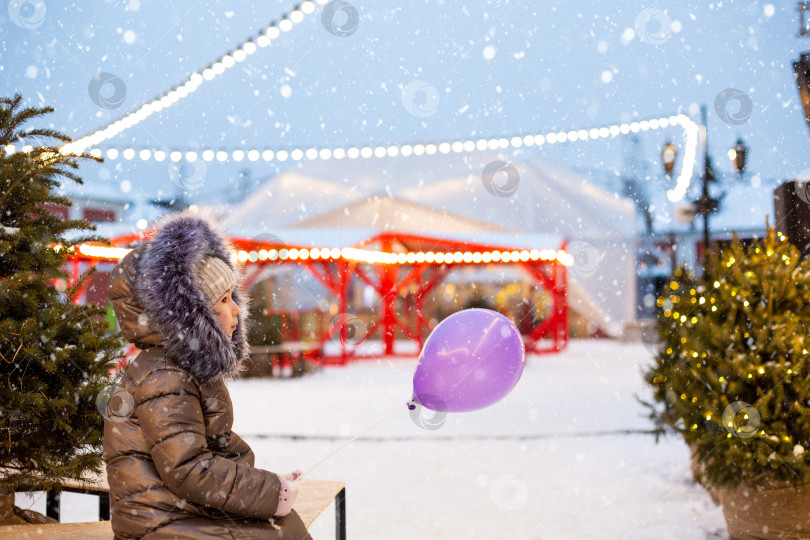 Скачать Девушка с рождественской елкой в руках на улице в теплой одежде зимой на праздничном базаре. Сказочные огни и гирлянды украсили снежный городок к новому году фотосток Ozero