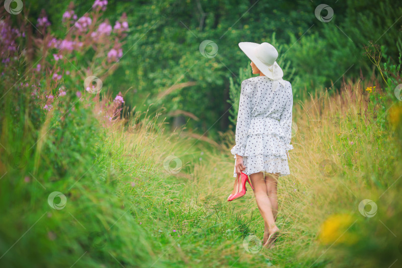 Скачать женщина в коротком белом платье и шляпке идет босиком по тропинке среди высокой травы с туфлями в руках фотосток Ozero