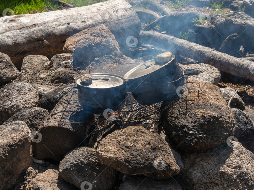 Скачать Походная кухня. Приготовление пищи на костре в дикой природе. Над огнем стоят два покрытых сажей чайника. Выживание в дикой природе. Чудесное пламя с кальдероном. Горшки в пламени костра. фотосток Ozero