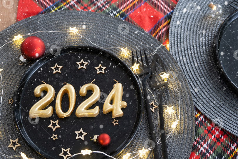 Скачать Золотые цифры 2024 года из свечей на черной тарелке на праздничном столе с новогодней сервировкой. Интерьер в стиле лофт, вечеринка, застолье. Плетеная салфетка, вилка, нож фотосток Ozero