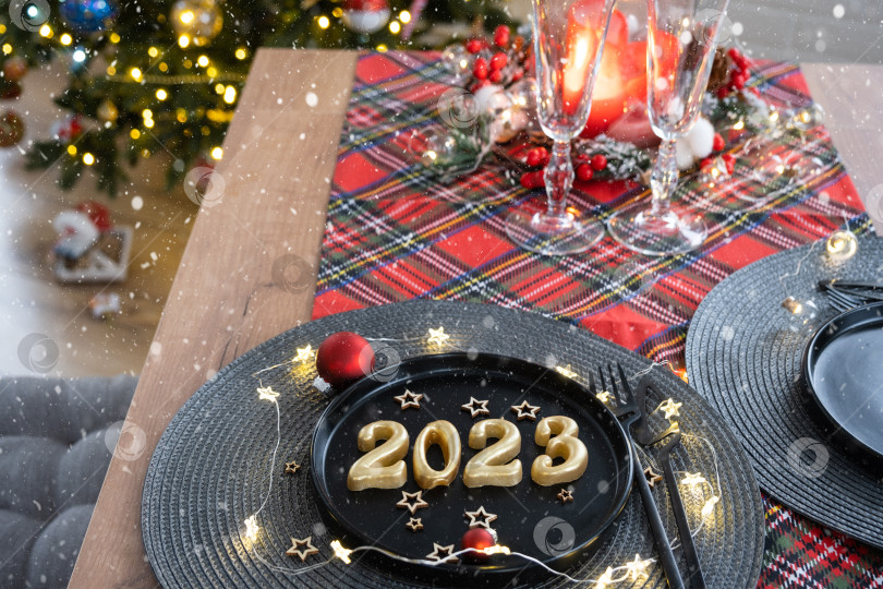 Скачать Золотые цифры 2023 года из свечей на черной тарелке на праздничном столе с новогодней сервировкой. Интерьер в стиле лофт, вечеринка, застолье. Плетеная салфетка, вилка, нож фотосток Ozero