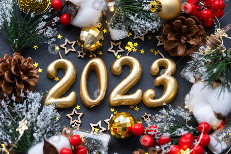 Скачать Золотые цифры 2023, выполненные из свечей на фоне черного каменного сланца, украшены праздничным декором из звезд, блесток, еловых веток, шаров и гирлянд. Поздравительная открытка, с Новым годом. фотосток Ozero