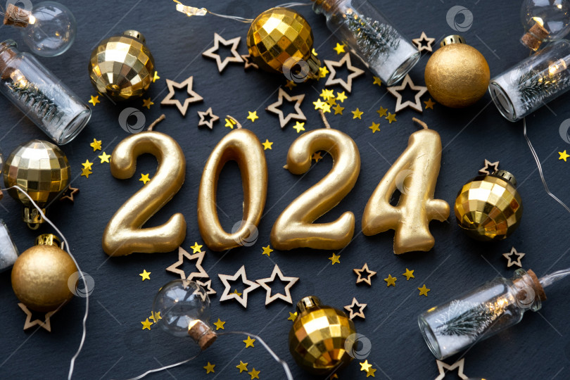 Скачать Золотые цифры 2024, выполненные из свечей на фоне черного каменного сланца, украшены праздничным декором из звезд, блесток, еловых веток, шаров и гирлянд. Поздравительная открытка, с Новым годом. фотосток Ozero