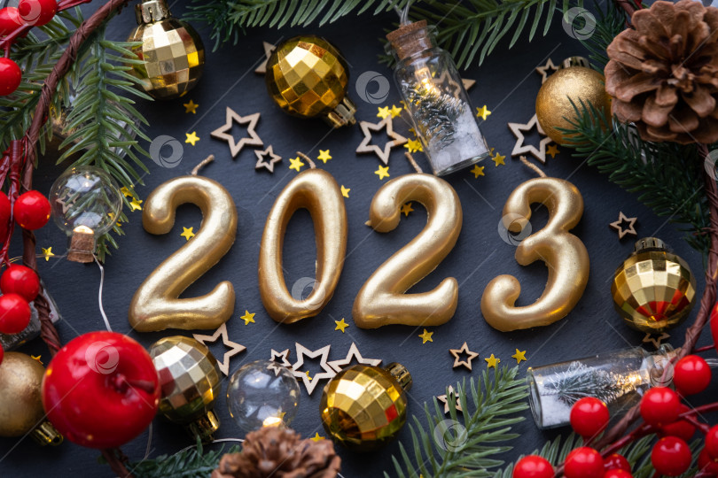Скачать Золотые фигуры 2023, выполненные из свечей на фоне черного каменного сланца, украшены праздничным декором из звезд, блесток, еловых веток, шаров и гирлянд. Поздравительная открытка с Новым годом. фотосток Ozero