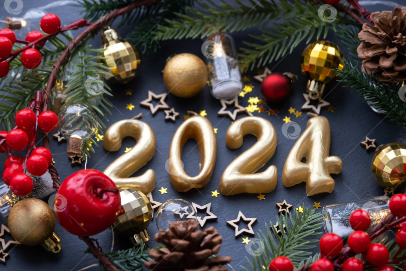 Скачать Золотые цифры 2024, выполненные из свечей на фоне черного каменного сланца, украшены праздничным декором из звезд, блесток, еловых веток, шаров и гирлянд. Поздравительная открытка с Новым годом. фотосток Ozero