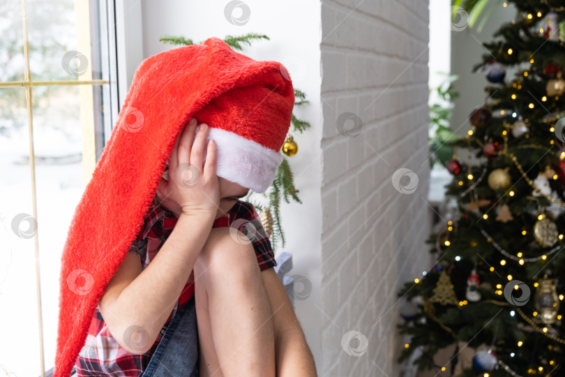 Скачать Несчастная девочка в шляпе Санта-Клауса сидит на подоконнике дома возле рождественской елки. Ребенок расстроен и плачет, ожидая Рождества и Нового года. Нет подарков, радости и чуда, разочарование фотосток Ozero