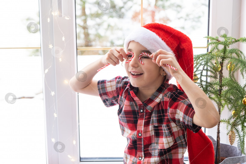 Скачать Девочка в шляпе Санта-Клауса сидит на подоконнике дома рядом с рождественской елкой и прикладывает леденцовую палочку к глазам, как очки. Ребенок веселится и корчит рожицы, ожидая Рождества и Нового года фотосток Ozero