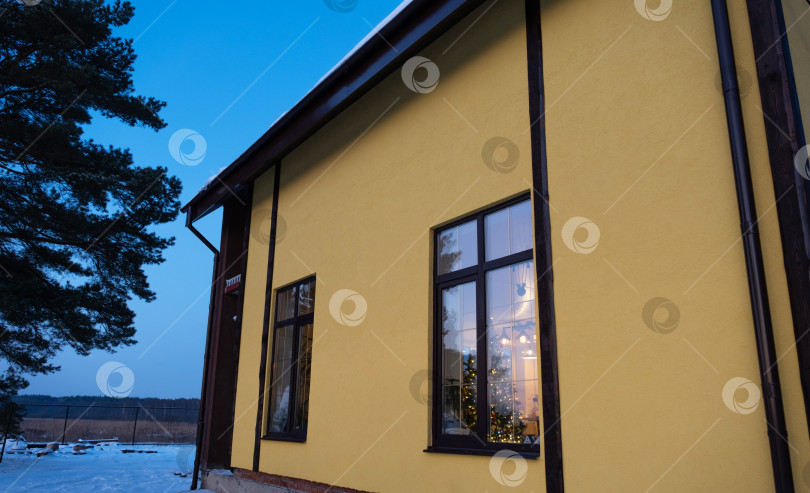 Скачать Зимой окна желтого дома выходят на улицу, покрытые снегом. Внутри жилища царит теплый свет, гирлянды гирлянд, рождественская елка. Уютное празднование Рождества и Нового года в домашних условиях фотосток Ozero