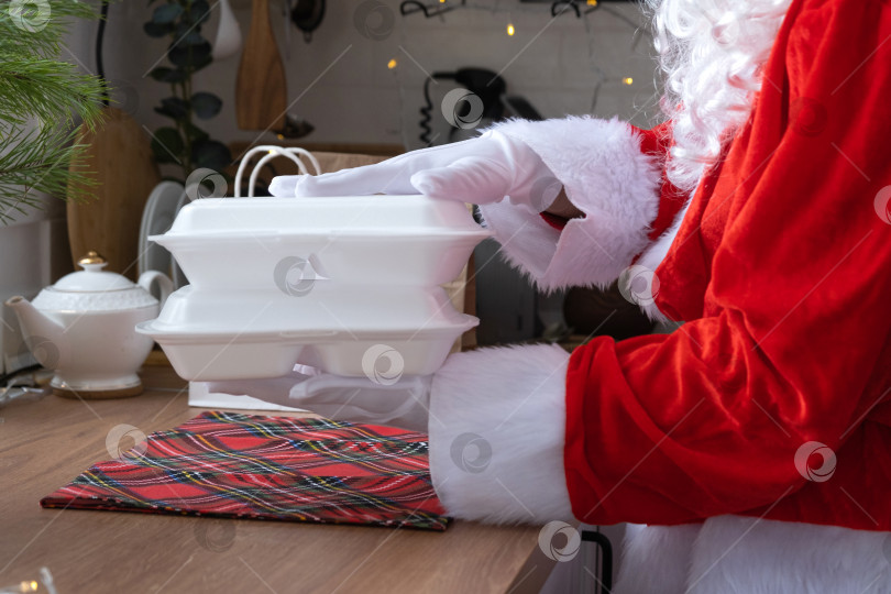 Скачать Сервис доставки еды на дом в контейнерах в руках Деда Мороза ставит их на кухонный стол и уносит. Готовый горячий заказ, кейтеринг на Рождественские, новогодние праздники. фотосток Ozero