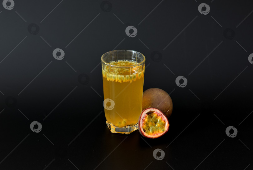Скачать Стакан сока из тропических фруктов с косточками на черном фоне рядом с кусочками спелой маракуйи. фотосток Ozero
