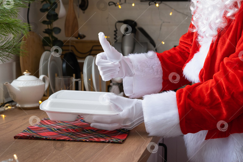 Скачать Сервис доставки еды на дом в контейнерах в руках Санта-Клауса ставит их на кухонный стол и забирает, подняв вверх большие пальцы. Готовый горячий заказ, кейтеринг на Рождественские, новогодние праздники. фотосток Ozero