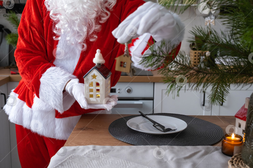 Скачать Ключ от дома с брелком-коттеджем в руках Санта-Клауса на открытом воздухе в снегу. Сделки с недвижимостью, покупка, строительство, переезд, ипотека. Уютный дом. Счастливого Рождества, новогоднего бронирования мероприятия и зала фотосток Ozero
