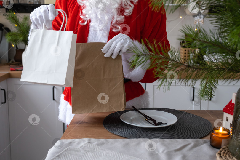 Скачать Санта-Клаус ставит его на кухонный стол и забирает бумажные пакеты с подарками для рукоделия, домашней выпечкой и доставкой еды. Покупки, переработка упаковки, ручная работа, доставка на Рождество и Новый год фотосток Ozero