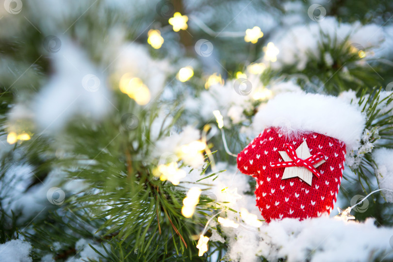 Скачать Красная рождественская игрушка в вязаных варежках на ветке натуральной ели, покрытой снегом. Новый год на открытом воздухе. Снегопад, огни гирлянд, праздничное настроение сказки и волшебства, уличное убранство. Пространство для копирования фотосток Ozero