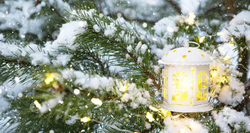 Скачать Рождественский фонарь с окнами на ветке натуральной ели в снегу. Новый год на открытом воздухе. Снегопад, огни гирлянд, праздничное настроение сказки и волшебства, уличное убранство. Пространство для копирования фотосток Ozero