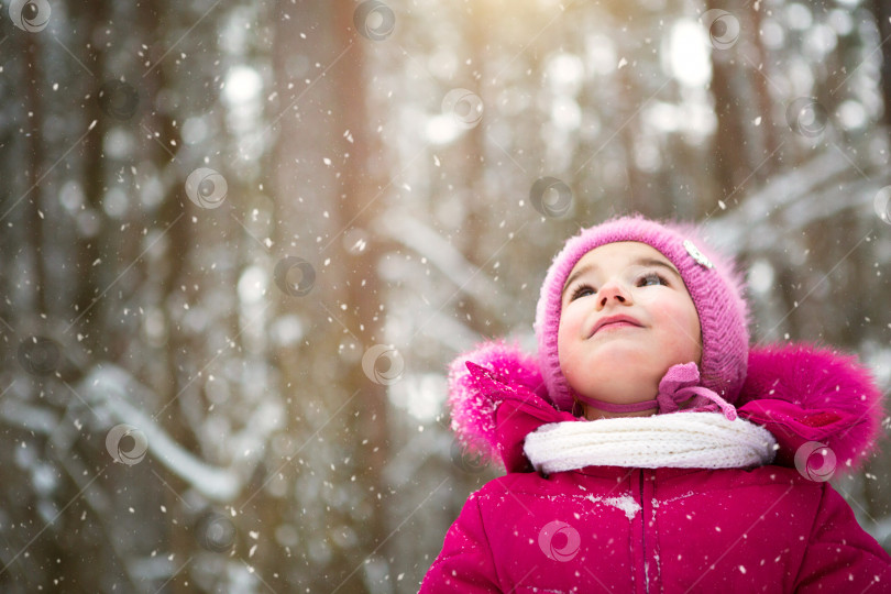 Скачать Маленькая девочка на морозе смотрит на снег с неба. Зима, гуляйте с малышом на свежем воздухе, снег. Теплая одежда, вязаная шапка, меховой помпон и капюшон. Игра в снежки, зимняя забава, фотосток Ozero