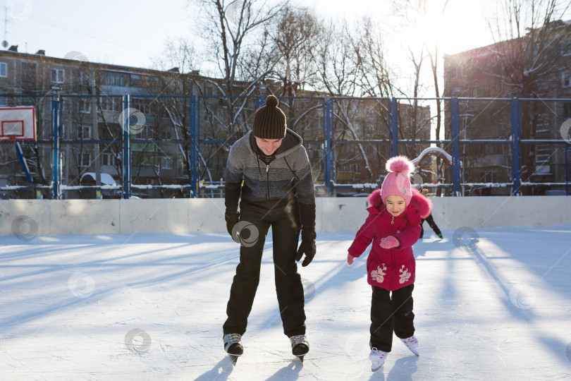 Скачать Папа учит свою маленькую дочь кататься на коньках на катке во дворе многоэтажных домов в городе. Морозный зимний солнечный день, активные зимние виды спорта и образ жизни фотосток Ozero