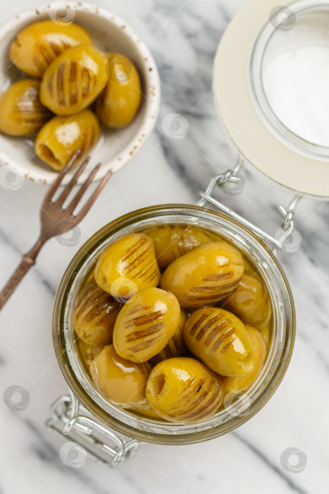 Скачать Зеленые оливки, приготовленные на гриле. Вкусная органическая оливка в стеклянной банке с оливковым маслом и чесноком на мраморном фоне. Выборочный фокус, вид сверху фотосток Ozero