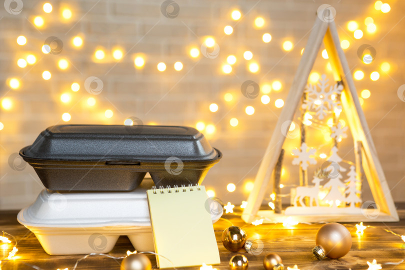 Скачать Рождественский декор контейнеров службы доставки еды. Акция в канун Нового года. Готовый горячий заказ, одноразовая пластиковая коробка в сказочном свете на столе. Работаем в праздничные дни в сфере общественного питания. Место для копирования, макет фотосток Ozero