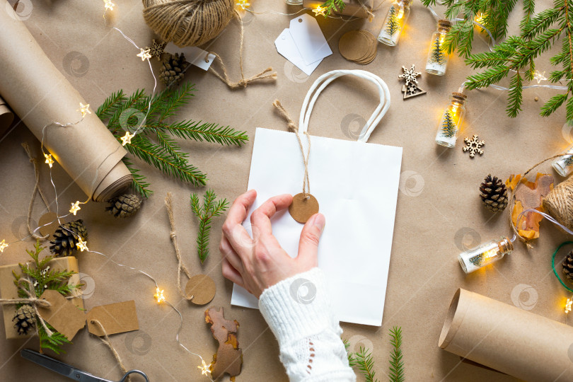 Скачать Руками в белом свитере упакуйте подарок на Рождество и новый год из экологически чистых материалов: крафт-бумаги, живых еловых веток, шишек, шпагата. Бирки с макетом, натуральный декор, ручная работа, сделай сам. Праздничное настроение фотосток Ozero