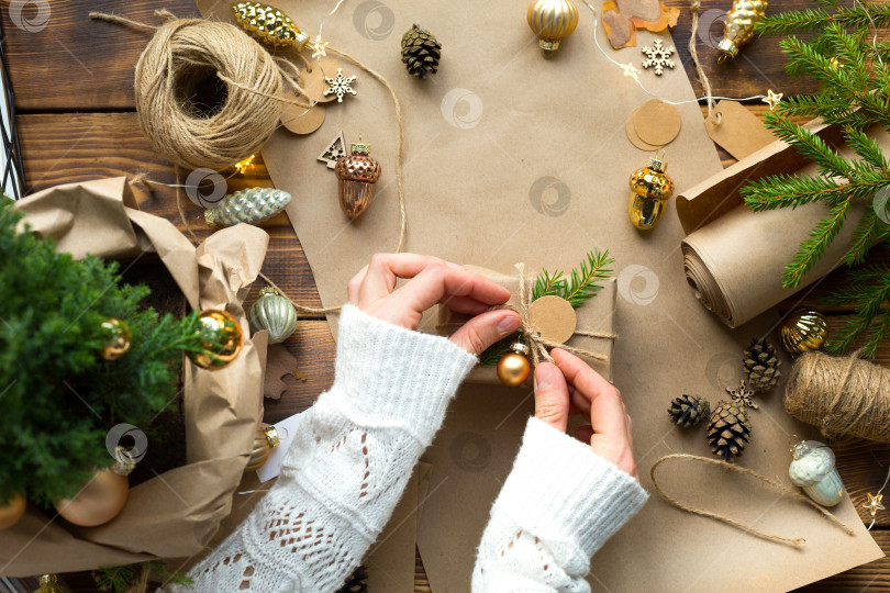 Скачать Руки в белом свитере упакуйте подарок на Рождество и новый год в экологически чистые материалы: крафт-бумагу, живые еловые ветки, шишки, шпагат. Бирки с макетом, натуральный декор, ручная работа, сделай сам. Праздничное настроение фотосток Ozero