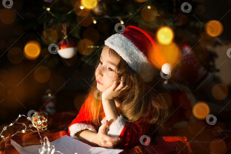 Скачать Маленькая девочка в шляпе Санты и красном платье под рождественской елкой мечтает, ожидая праздника, лежа на клетчатом одеяле. Письмо на клочке бумаги, подарки. Новый год, Рождество. расфокусировка света фотосток Ozero