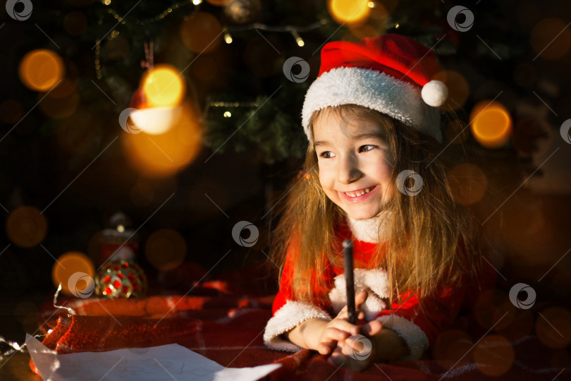 Скачать Маленькая девочка в шляпе Санты и красном платье под рождественской елкой мечтает, ожидая праздника, лежа на клетчатом одеяле. Письмо на клочке бумаги, подарки. Новый год, Рождество. расфокусировка света фотосток Ozero