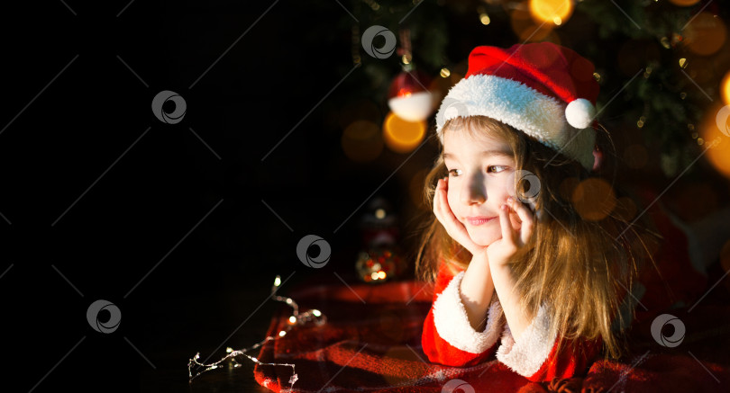 Скачать Маленькая девочка в шляпе Санты и красном платье под рождественской елкой мечтает, ожидая праздника, лежа на клетчатом одеяле. Письмо на листе бумаги, подарки. Новый год, Рождество. расфокусировка света фотосток Ozero