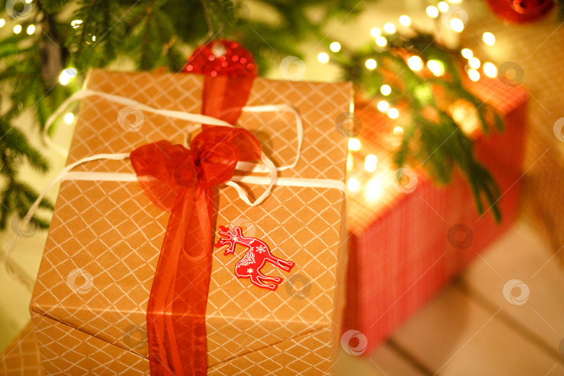 Скачать Подарочная коробка в золотистой клетчатой обертке перевязана красным бантом под рождественской елкой. Рождество, новый год, гирлянды, подвеска с оленем. Пространство для текста фотосток Ozero