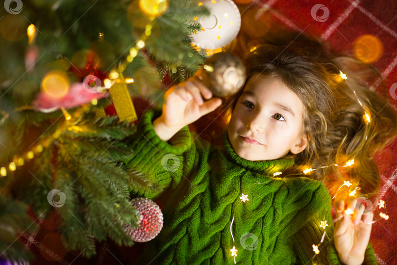 Скачать Девочка с длинными волосами и гирляндами лежит на красном пледе под рождественской елкой с игрушками в теплом вязаном свитере. Рождество, Новый год, детские эмоции, радость, ожидание чуда и подарков фотосток Ozero