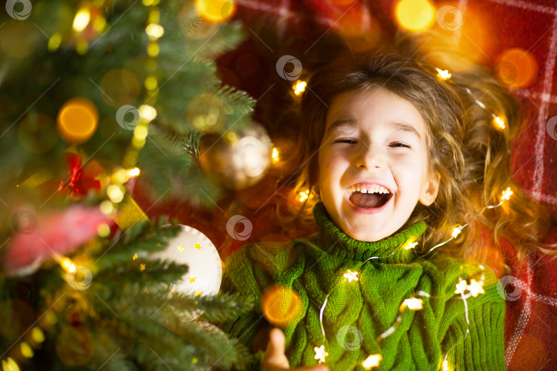 Скачать Девочка с длинными волосами и гирляндами лежит на красном пледе под рождественской елкой с игрушками в теплом вязаном свитере. Рождество, Новый год, детские эмоции, радость, ожидание чуда и подарков фотосток Ozero