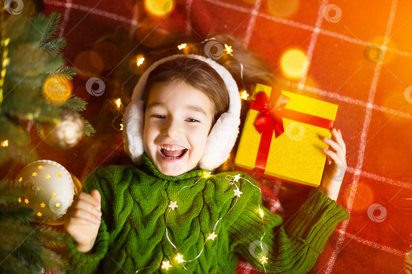 Скачать Девушка в теплом вязаном свитере и меховых наушниках лежит на одеяле возле рождественской елки с подарочной коробкой. Канун Рождества, мечта и желание. Новый год, праздничная атмосфера, гирлянды и боке фотосток Ozero
