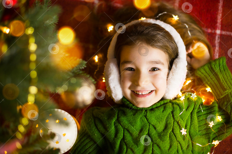 Скачать Счастливая девушка в меховых наушниках лежит возле рождественской елки в гирляндах и огоньках, в теплом свитере на уютном одеяле. Рождество и Новый год, канун праздника, волшебное настроение фотосток Ozero