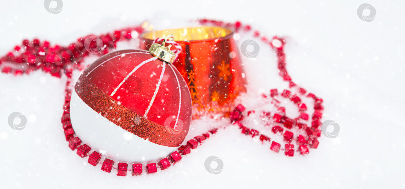 Скачать Красный рождественский шар на натуральном белом снегу с квадратными бусинами и горящей свечой в стеклянном подсвечнике. Рождество, Новый год на открытом воздухе. Снегопад, праздничное настроение сказки и волшебства, уличное убранство. фотосток Ozero