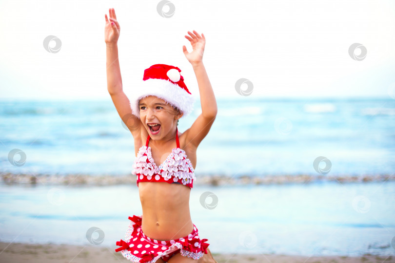 Скачать 5-летняя девочка в красном купальнике и шляпе Санты на пляже счастлива, кричит, прыгает и машет руками от счастья. Рождественские и новогодние зимние туры в теплые страны. Туризм для отдыха в тропиках фотосток Ozero