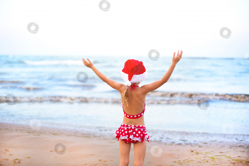 Скачать 5-летняя девочка в красном купальнике и шляпе Санты на пляже счастлива, кричит, прыгает и машет руками от счастья. Рождественские и новогодние зимние туры в теплые страны. Туризм для отдыха в тропиках фотосток Ozero