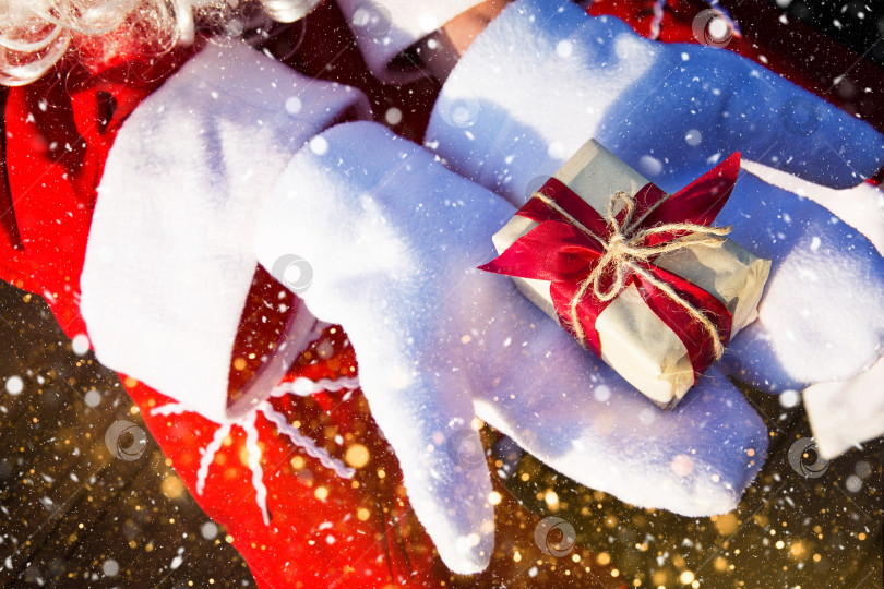Скачать Коробка с рождественским подарком в руках Деда Мороза в белых варежках. Красный костюм, борода, гирлянды огней - все как в тумане. Новый год, подготовка, ожидание чуда, сбывшейся мечты. Закрыть фотосток Ozero