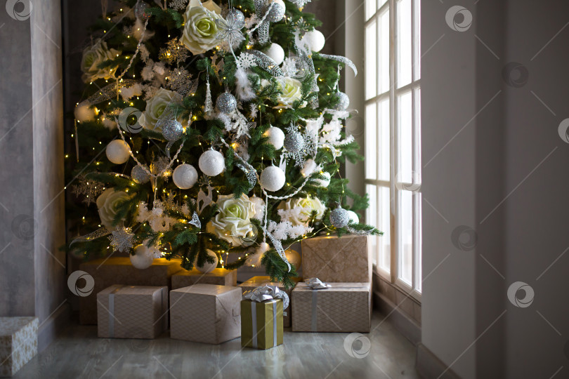 Скачать Подарочная коробка в золотой упаковке и серебряный бант под рождественской елкой с белым декором. Новый год, сочельник, поздравления для семьи, уют и ожидание чуда фотосток Ozero
