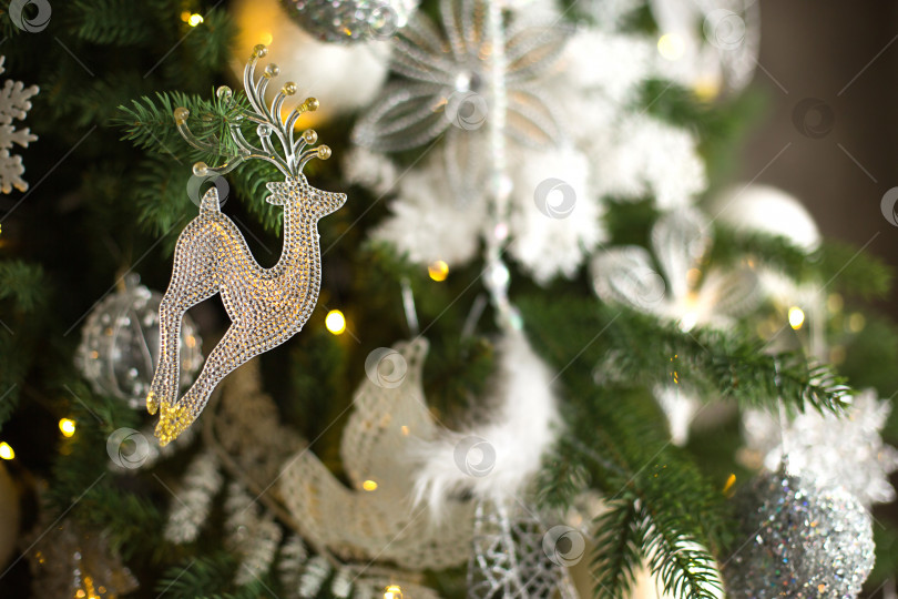 Скачать Подвеска в виде оленя из прозрачного стекла на рождественской елке с гирляндами огней. Белый рождественский декор, перья, воздушные шары, цветы, ленты. Новый год, атмосфера праздника и волшебства. Пространство для текста фотосток Ozero