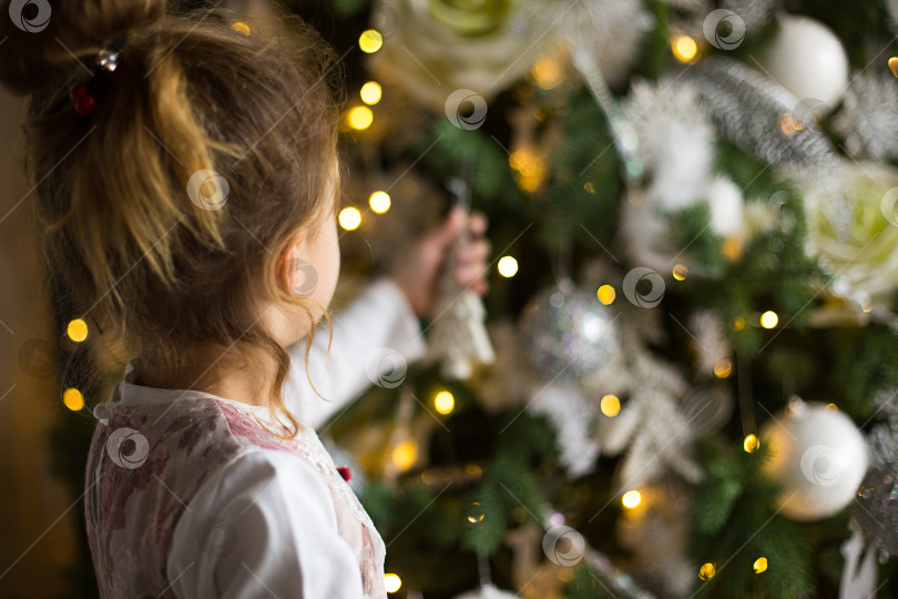 Скачать Маленькая девочка тянется рукой к рождественской елочной игрушке. Рождественский декор в ожидании праздника и чуда. новый год. Белый, розовый и голубой декор, золотистые гирлянды фотосток Ozero