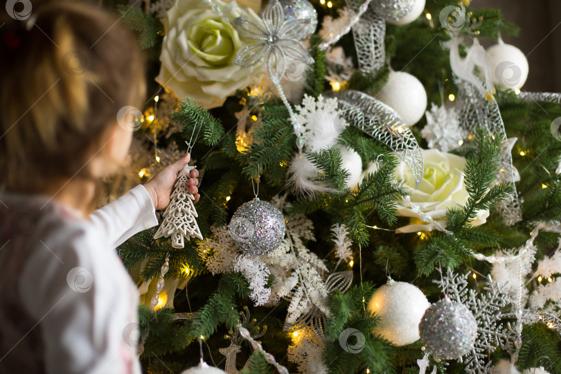 Скачать Маленькая девочка тянется рукой к рождественской елочной игрушке. Рождественский декор в ожидании праздника и чуда. новый год. Белый, розовый и голубой декор, золотые гирлянды фотосток Ozero