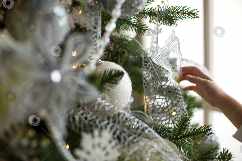 Скачать Подвеска в виде оленя из прозрачного стекла на рождественской елке с гирляндами огней. Белый рождественский декор, перья, воздушные шары, цветы, ленты. Новый год, атмосфера праздника и волшебства. Пространство для текста фотосток Ozero