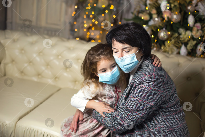 Скачать Бабушка и внучка на диване в гостиной с рождественским декором обнимаются в медицинских масках на лицах. Семейный отдых во время вспышки коронавируса и других заболеваний. новый год фотосток Ozero