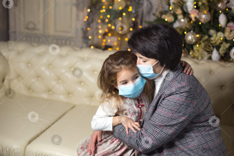 Скачать Бабушка и внучка на диване в гостиной с рождественским декором обнимаются в медицинских масках на лицах. Семейный отдых во время вспышки коронавируса и других заболеваний. новый год фотосток Ozero