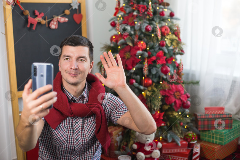 Скачать Молодой человек возле рождественской елки у себя дома в удобной одежде машет рукой в знак приветствия / прощания перед экраном смартфона. Поздравляем по видеосвязи с Рождеством и новым годом. фотосток Ozero